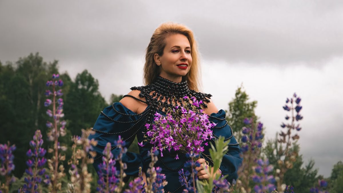 A woman in a blue dress with purple flowers stands in a lush lupine field under cloudy skies.